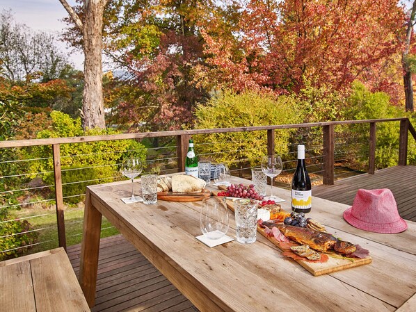 A wooden deck with a table set for a meal, featuring a bottle of wine, glasses, bread, and a charcuterie board.