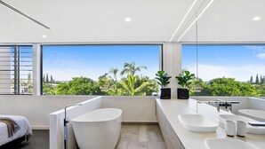 A modern bathroom with a bathtub, two sinks, and a large window offering a view of greenery and buildings. - Blueys Beach House - 4 bedroom (Sunrise Beach)