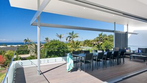 A modern outdoor patio with a view of the ocean, featuring a dining area with tables and chairs, a seating area with a sofa, and a wooden deck. - Blueys Beach House - 4 bedroom (Sunrise Beach)