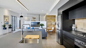 A modern kitchen with a built-in stove, a large island with a sink, and a dining area with a view of the beach. - Blueys Beach House - 4 bedroom (Sunrise Beach)