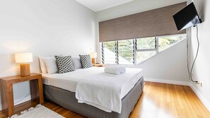 Un dormitorio moderno con cama, mesita de noche con lámpara, ventana con persianas y televisor montado en la pared.