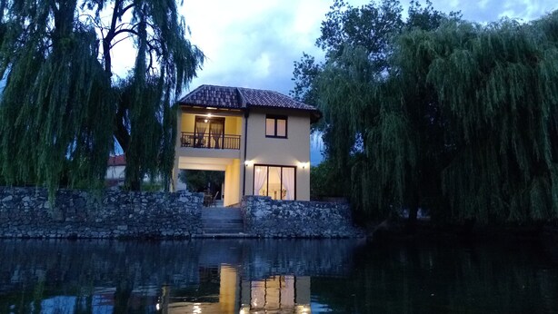 Una casa de dos pisos con un balcón, un muro de piedra y un reflejo en el agua.