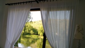 Habitación con un ventanal grande, cortinas blancas, vista a un estanque y una lámpara sobre una mesita de noche.