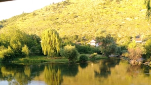 Una mesa con un farol metálico sobre un posavasos de encaje, con vistas a un río tranquilo que refleja árboles y una colina al fondo.