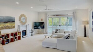 A modern living room with a white sofa, a flat-screen TV, a fireplace, and a large window with curtains. - Tropical Family Oasis, Sunshine Beach (Sunshine Beach)