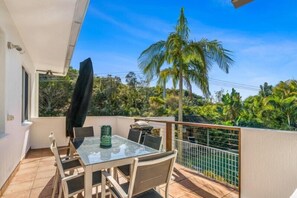 A balcony with a glass table and chairs, a grill, and a view of palm trees and greenery.