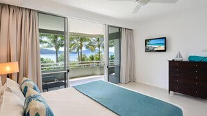 A bedroom with a bed, a dresser, and a view of palm trees and the ocean. - Hibiscus 107 - Hamilton Island (Whitsundays)
