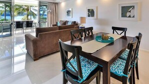 A modern dining area with a wooden table and chairs, a brown sofa, and a view of the ocean through large windows. - Hibiscus 107 - Hamilton Island (Whitsundays)