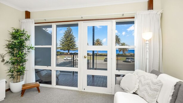 A living room with a large sliding glass door leading to a balcony, a white sofa, a wooden bench, and a potted plant. - Beautiful Beach Front Townhouse (Victor Harbor)
