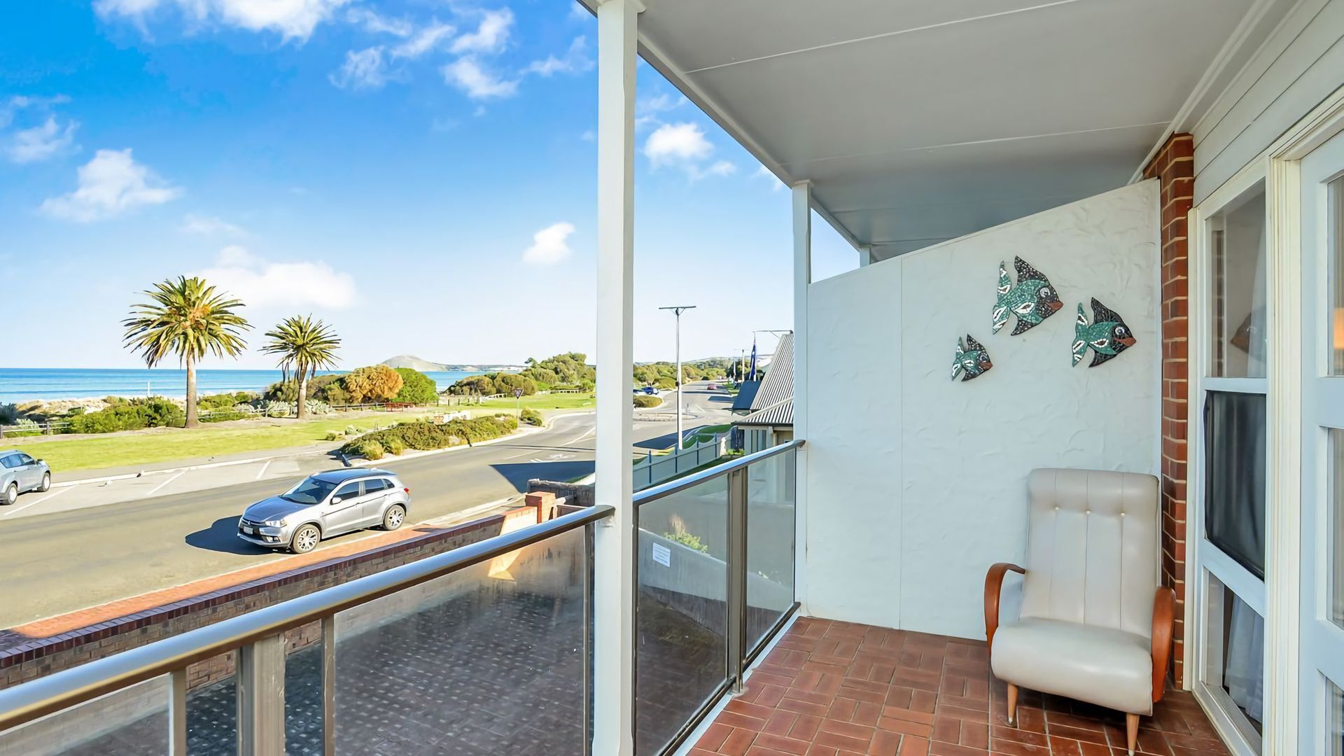 A balcony with a view of a road, palm trees, and a building with decorative fish wall art.