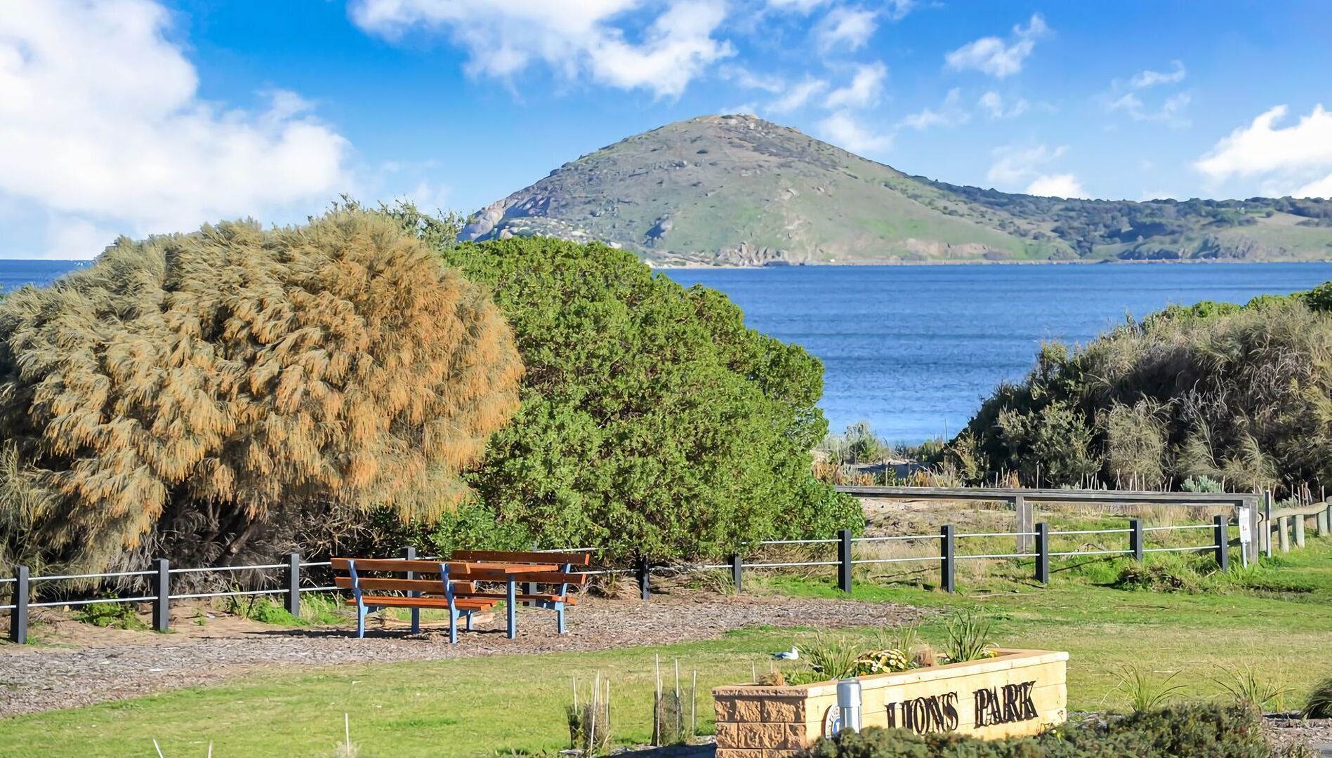 A park with a bench, a sign reading "Lions Park," and a view of the sea and a hill in the background.