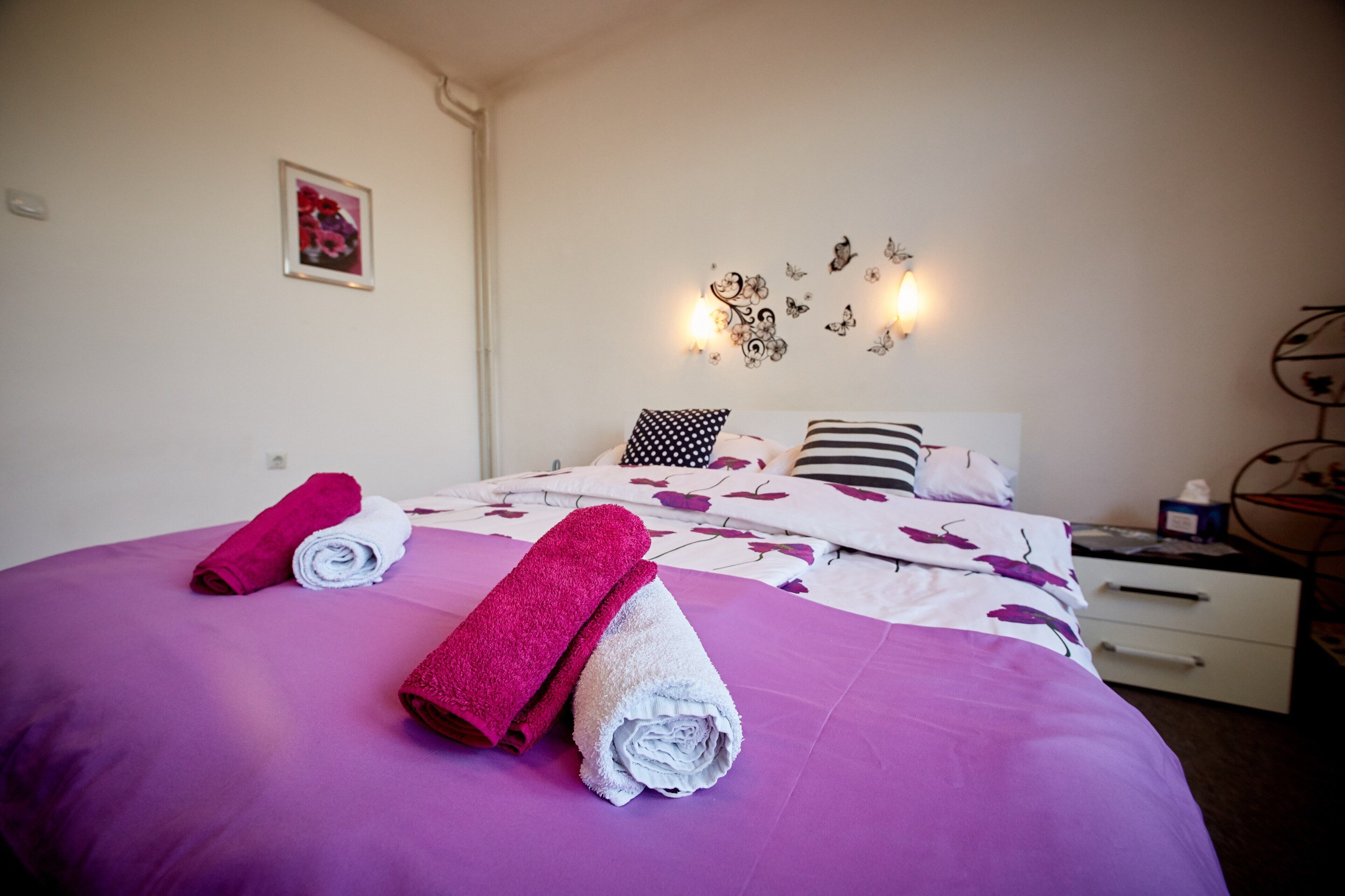 Ein ordentlich bezogenes Bett mit rosa und weißen Handtüchern, einer floralen Tagesdecke und einer wandmontierten Leuchte mit dekorativen Elementen.