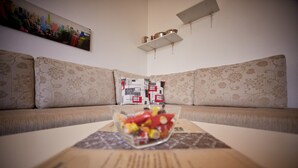 Een woonkamer met een bank, een bijzettafeltje met een schaal fruit en een plank met boeken en decoratieve objecten.
