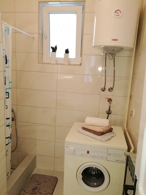 A compact bathroom with a washing machine, a small window with plants, and a shower area.