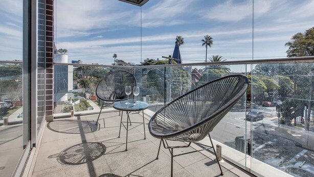A balcony with two wicker chairs and a small table, overlooking a cityscape with palm trees and a church steeple.