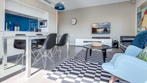 A modern living room with a checkered floor, a blue sofa, a dining area with a white table and chairs, a kitchen with a blue backsplash, and a clock on the wall.