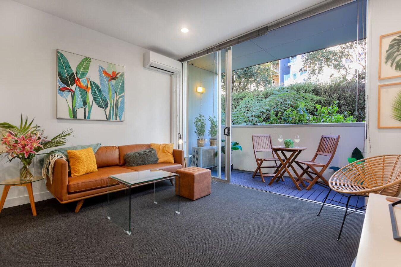 A modern living room with a sofa, coffee table, and chairs, a glass door leading to a balcony with plants and a view of greenery.