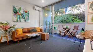 Un salón moderno con sofá, mesa de centro y sillas, puerta de cristal que da a un balcón con plantas y vistas al verdor.