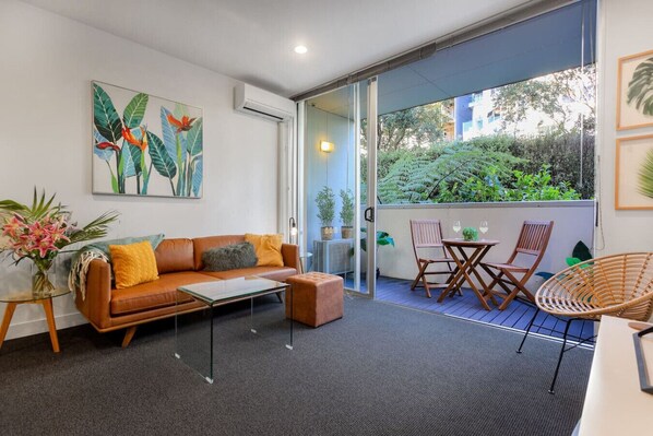 A modern living room with a sofa, coffee table, and chairs, a glass door leading to a balcony with plants and a view of greenery.