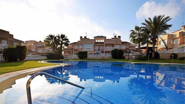 A swimming pool with clear blue water, surrounded by palm trees and a building with balconies in the background. - Cosy flat on second floor with large pool (Orihuela)