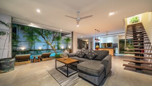 Uma sala de estar moderna com um sofá cinza, mesa de centro de madeira e cadeiras de vime. Um grande mural com árvores tropicais e uma piscina está na parede.