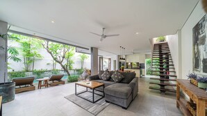 Uma sala de estar ampla com sofá cinzento, mesa de centro de madeira e mobiliário de vime. Uma escada com guarda de plantas leva a um andar superior.