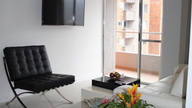 A modern living room with a black leather chair, a white sofa, a flat-screen TV mounted on the wall, and a balcony with a view of a brick building.