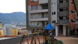 A playground with swings and climbing structures, surrounded by a brick building with multiple balconies.