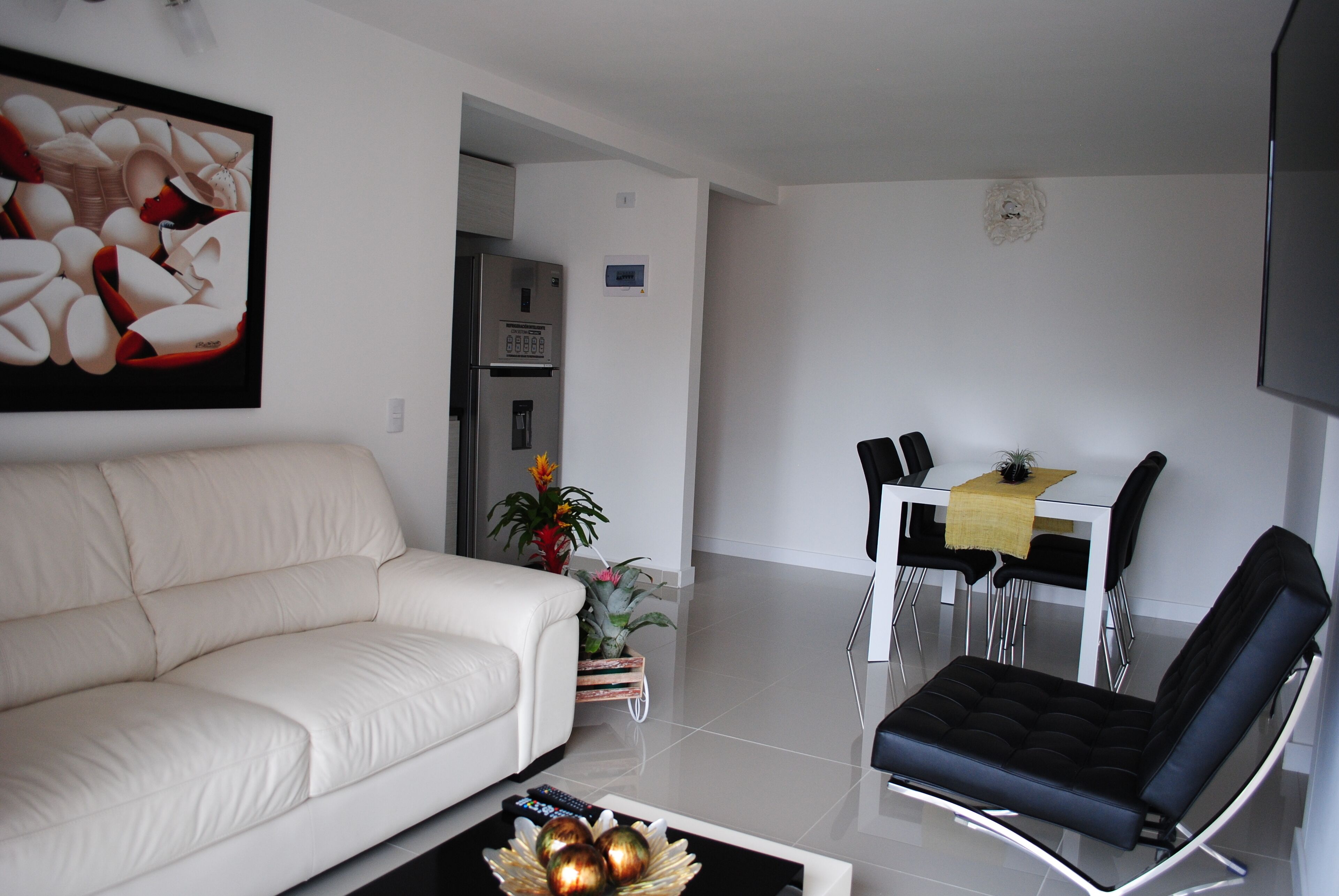 A modern living room with a white sofa, a black armchair, a dining table with chairs, and a refrigerator.