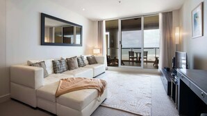 A modern hotel room with a sofa, a flat-screen TV, a large window, and a sliding glass door leading to a balcony. - Ocean View 1BR Private balcony OAKS PIER (Glenelg)