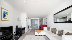 A modern living room with a flat-screen TV, a sofa, a coffee table, and a large mirror. - Ocean View 1BR Private balcony OAKS PIER (Glenelg)