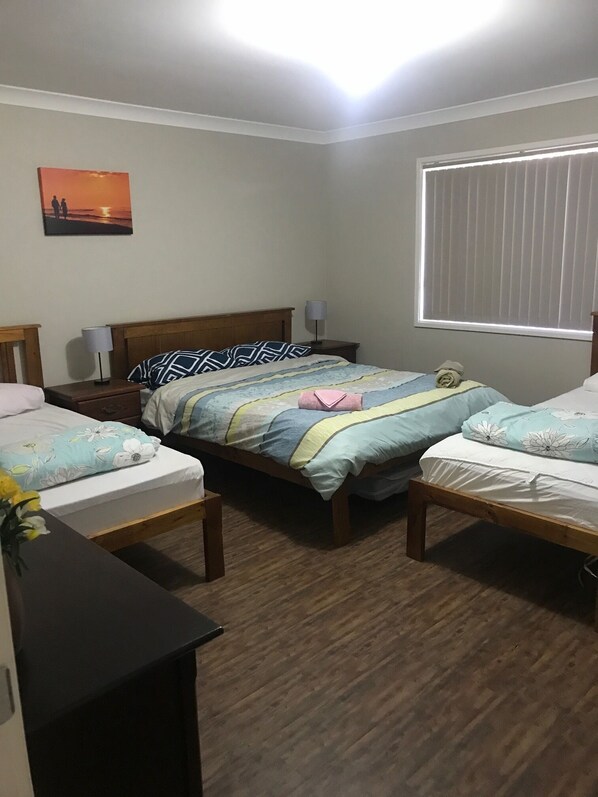 Una habitación con dos camas individuales, cabecero de madera, mesita de noche con lámpara, un cuadro colgado en la pared y una ventana con persianas.