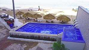 A pool with a blue mosaic tile interior, surrounded by lounge chairs and thatched umbrellas, overlooking a beach. - Beachfront right on Los Muertos beach (Amapas)