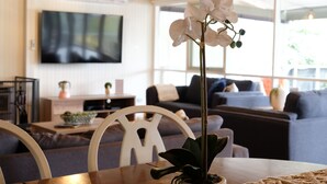 A modern living room with a large flat-screen TV mounted on the wall, a dining table with a potted orchid, and a sofa set in the background.