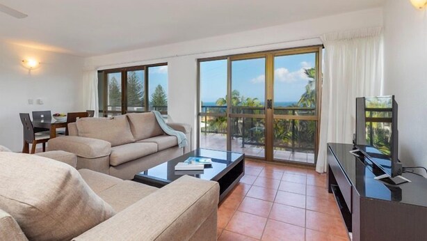 Una sala de estar moderna con sofá, mesa de centro y televisor, con puertas corredizas de vidrio que dan a un balcón con vista al océano.