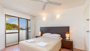 Un dormitorio con una cama, dos mesitas de noche con lámparas, un ventilador de techo y un ventanal corredero que da a un balcón.