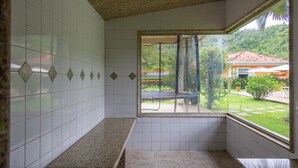 Un baño con paredes de azulejos blancos, un banco y una ventana con vistas a una casa y vegetación.