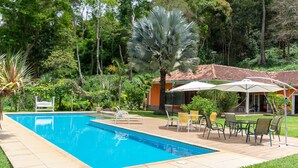 Una piscina con solárium empedrado, rodeada de exuberante vegetación y un edificio con tejado de azulejos.