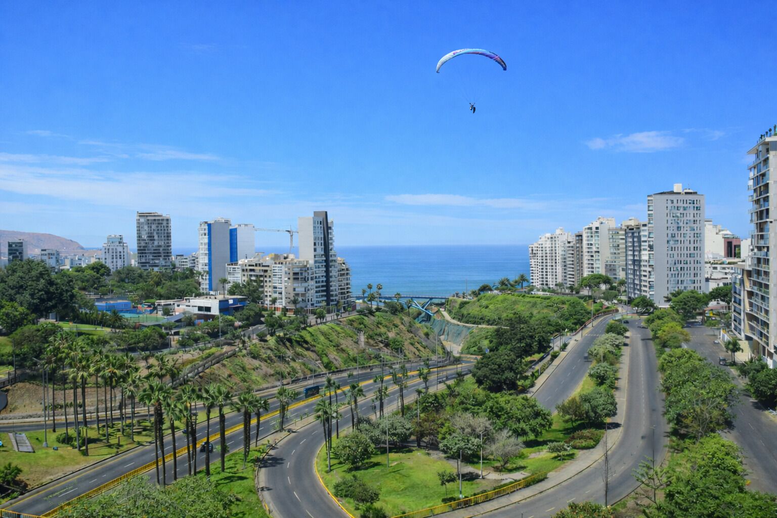 A coastal cityscape with modern buildings, a winding road, and a person paragliding.