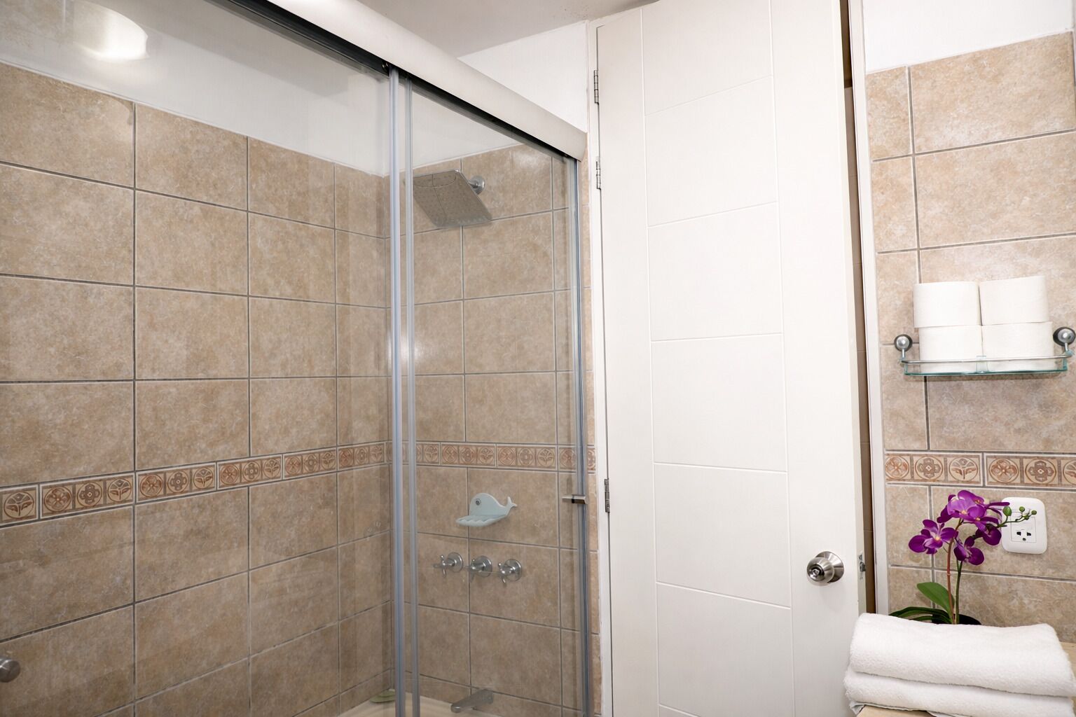 Un baño con azulejos color beige, cabina de ducha de vidrio, toallero blanco y una orquídea púrpura.