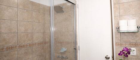 Un baño con azulejos color beige, cabina de ducha de vidrio, toallero blanco y una orquídea púrpura.