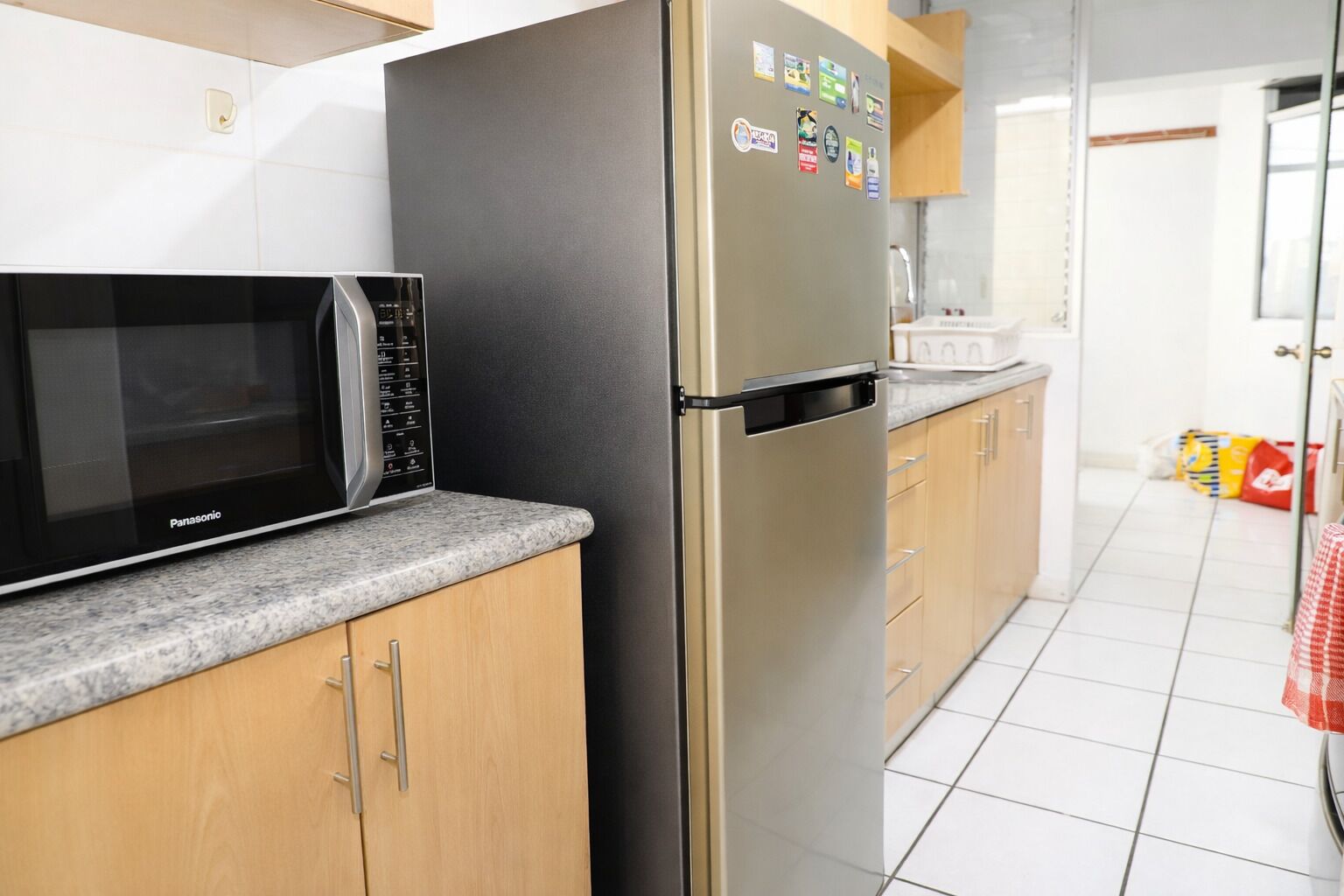 A kitchen with a black microwave, a stainless steel refrigerator, wooden cabinets, and a tiled floor.