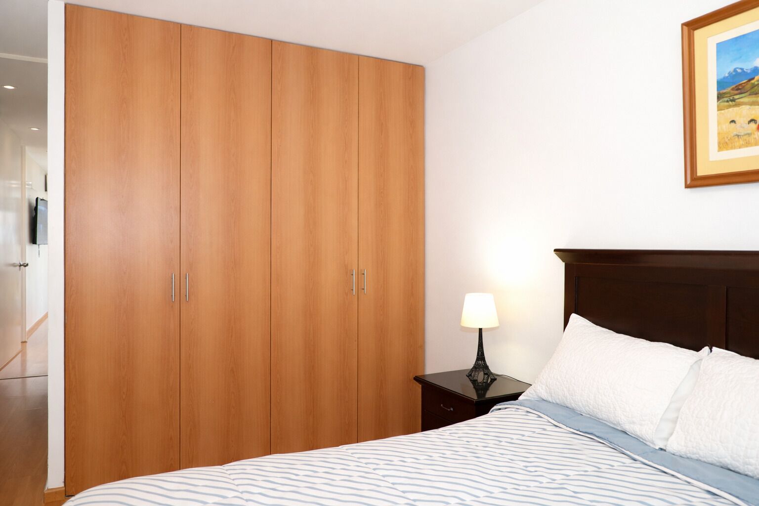 A bedroom with a bed, a nightstand with a lamp, a wooden wardrobe, and a framed picture on the wall.