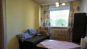Une petite pièce agréable et chaleureuse, avec un lit, une table et une chaise. Il y a une fenêtre avec des rideaux à motifs floraux et un radiateur en dessous.