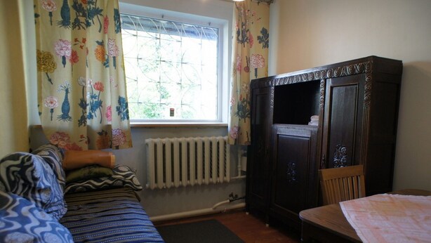 Une pièce comprenant un lit, une armoire en bois, un radiateur et une fenêtre ornée de rideaux à motifs floraux.
