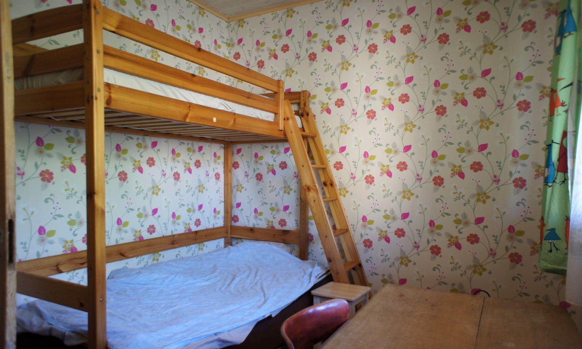 Une chambre avec un lit superposé, un papier peint à motifs floraux, une échelle et une petite table en bois.