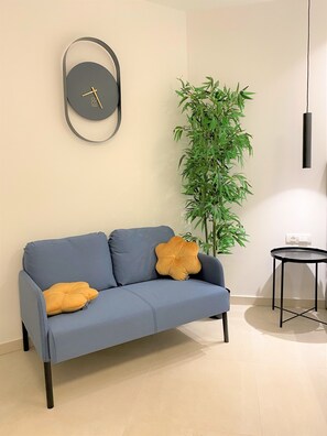 Un salon moderne avec un canapé bleu, deux coussins de couleur moutarde, une horloge murale ronde, une grande plante verte, une petite table ronde et un luminaire suspendu.