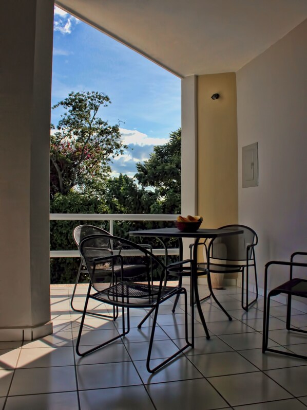 Un balcon avec une table et des chaises, un saladier rempli de fruits et une vue sur les arbres et le ciel.