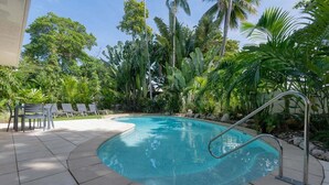 Una piscina con una escalera metálica, rodeada de plantas y árboles tropicales.