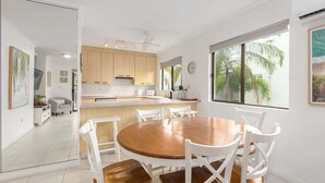 Una cocina moderna con armarios blancos, una mesa de comedor de madera y sillas. Se observa un ventanal con vistas a palmeras.